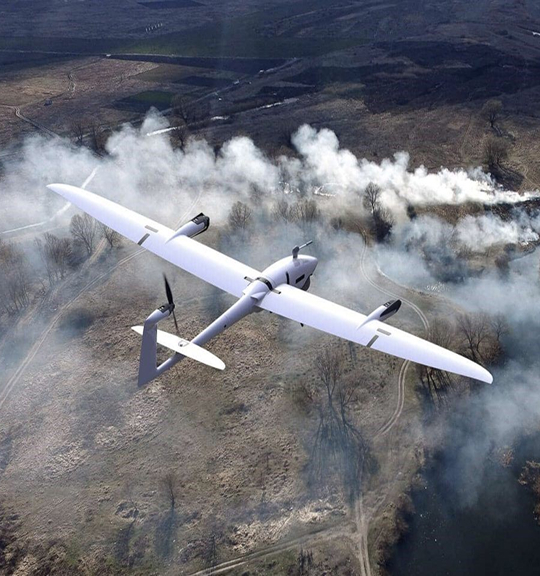 Quantum Systems Vector Drone Flight A Quantum Systems Vector fixed-wing drone in mid-flight over a rugged, smoky terrain. The UAV, equipped with front and rear propellers and visible sensor pods, soars above a landscape marked by plumes of smoke rising from the ground.
